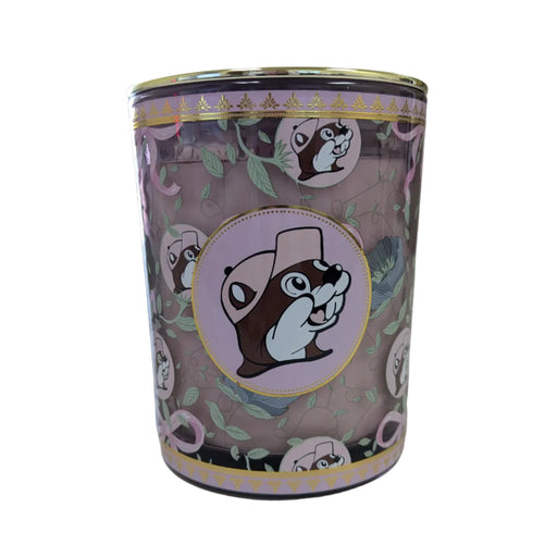 A close-up shot of a Buc-ee’s Blossoms & Bows scented candle in a purple glass jar with a gold lid. The decorative design features a repeating motif of pink bows and green leaves. The central logo shows Buc-ee the Beaver in a pink hat on a light pink circular backdrop with a gold-leaf border. The candle wax is a soft mauve-purple color. Displayed on a clean white background.