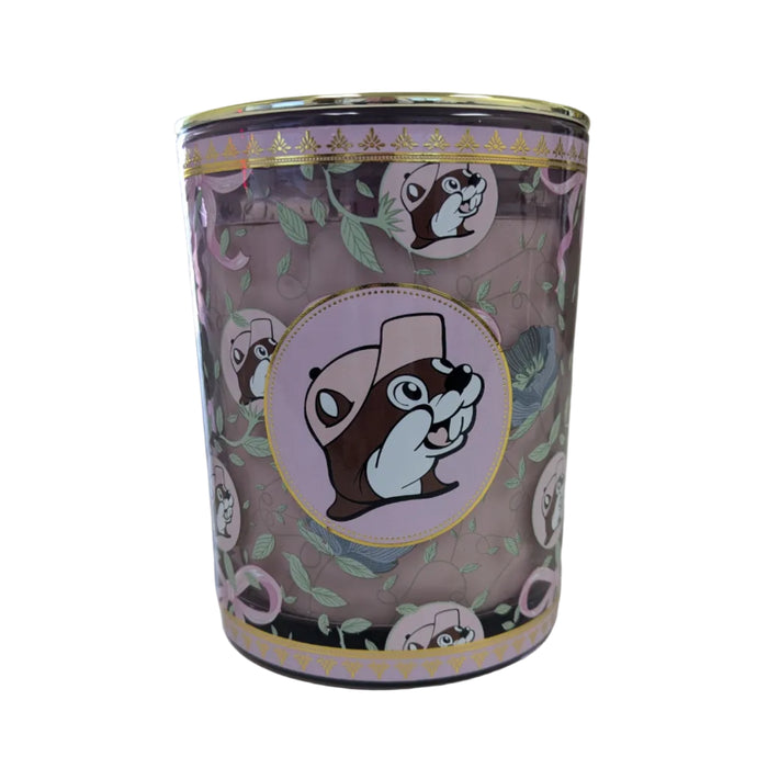 A close-up shot of a Buc-ee’s Blossoms & Bows scented candle in a purple glass jar with a gold lid. The decorative design features a repeating motif of pink bows and green leaves. The central logo shows Buc-ee the Beaver in a pink hat on a light pink circular backdrop with a gold-leaf border. The candle wax is a soft mauve-purple color. Displayed on a clean white background.