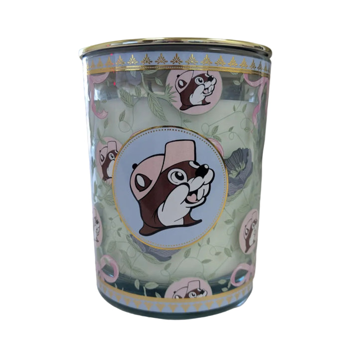 A close-up shot of a Buc-ee’s Blossoms & Bows scented candle in a teal glass jar with a gold lid. The jar is decorated with a pattern of green vines, purple flowers, and pink bows. A central circular emblem features the Buc-ee the Beaver mascot wearing a pink hat against a light blue background, bordered by a gold rim. The wax inside is a pale, off-white color. Displayed on a clean white background.
