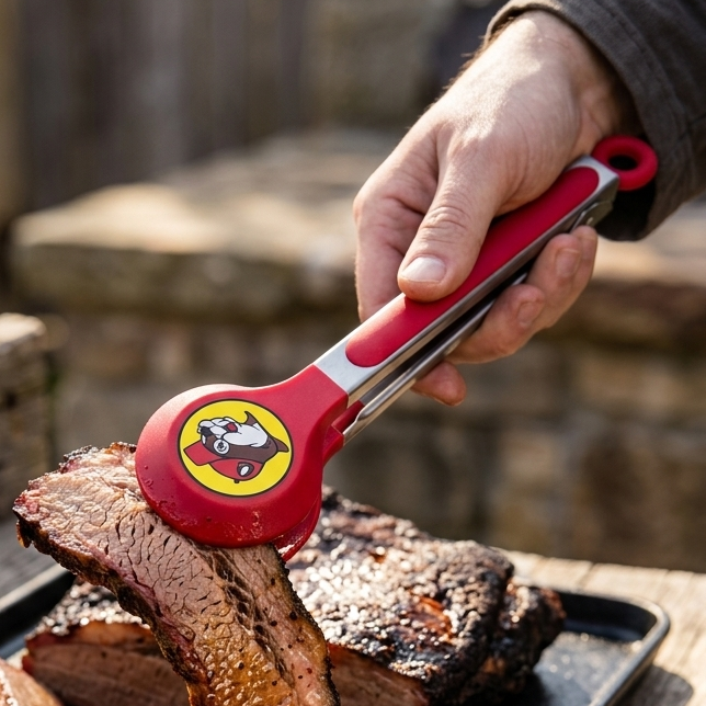 A lifestyle photo of a person using Buc-ee's red and stainless steel serving tongs with a beaver logo to lift a slice of sliced brisket from a black tray on a wooden table.