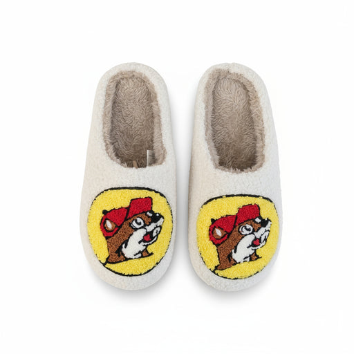 Pair of white-colored fuzzy slippers featuring the Buc-ee’s beaver logo on the front in red, yellow, brown, and white. The slippers have a soft sherpa-like interior and plush exterior, displayed on a plain white background.