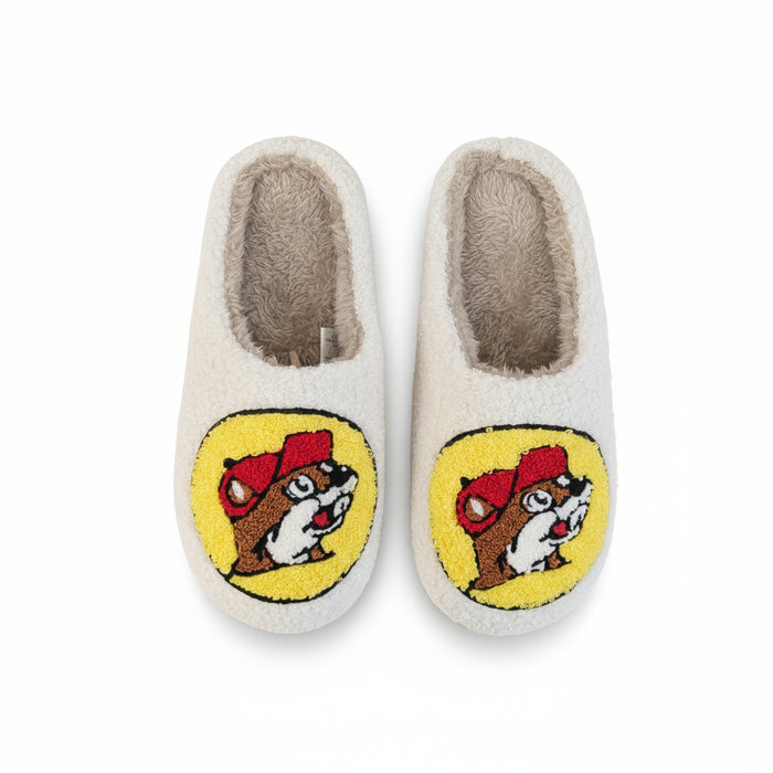 Pair of white-colored fuzzy slippers featuring the Buc-ee’s beaver logo on the front in red, yellow, brown, and white. The slippers have a soft sherpa-like interior and plush exterior, displayed on a plain white background.