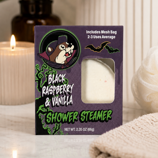Black Raspberry & Vanilla Shower Steamer in its purple Halloween-themed box displayed on a white marble bathroom countertop with a lit candle, rolled towel, and greenery in the background, highlighting the product in a cozy spa-inspired setting.