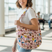 A lifestyle photo of a young woman with short brown hair smiling and looking over her shoulder while walking through a bright airport terminal. She is wearing a white t-shirt and blue jeans, carrying the Buc-ee’s Snacks Pink Puffy Tote Bag on her left shoulder. The bag's light pink, quilted fabric and snack-themed print are prominently displayed, including the front zippered pocket with pink pull strings. 