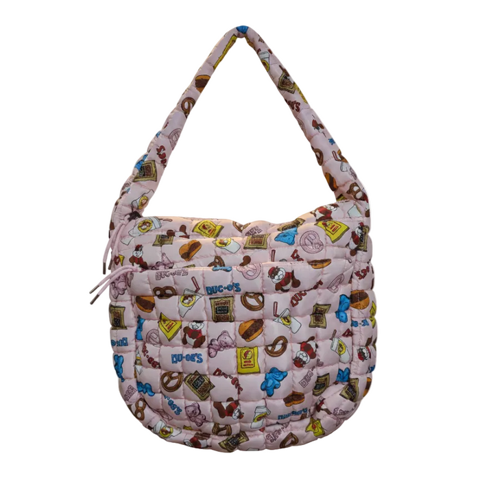 A professional product shot of the Buc-ee’s  Snacks Pink Puffy Tote Bag isolated against a solid white background. The tote features a light pink, quilted "puffy" exterior decorated with a colorful all-over print of Beaver Nuggets, pretzels, gummy bears, and soda cups. The bag is shown with its thick, matching quilted shoulder strap arched upward as if being carried. On the front, a horizontal zippered pocket is visible, accented with two pink pull strings.