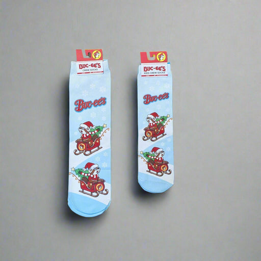 Two pairs of Buc-ee’s Snowflake Christmas Crew Socks, one kids’ size and one adult size, featuring a light blue background with snowflakes and Buc-ee riding in a Christmas sleigh filled with presents.