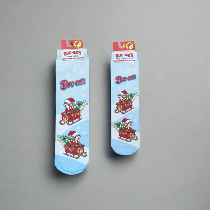 Two pairs of Buc-ee’s Snowflake Christmas Crew Socks, one kids’ size and one adult size, featuring a light blue background with snowflakes and Buc-ee riding in a Christmas sleigh filled with presents.