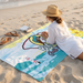 A lifestyle photograph from a high angle showing a woman wearing a white beach cover-up and straw hat, lying on her stomach on a large beach towel spread over the sand. The towel prominently displays the custom Buc-ee’s South Carolina coastal design, featuring a white lighthouse with a pink roof, the iconic beaver mascot, and yellow flowers, with ocean waves washing onto the shore in the background.