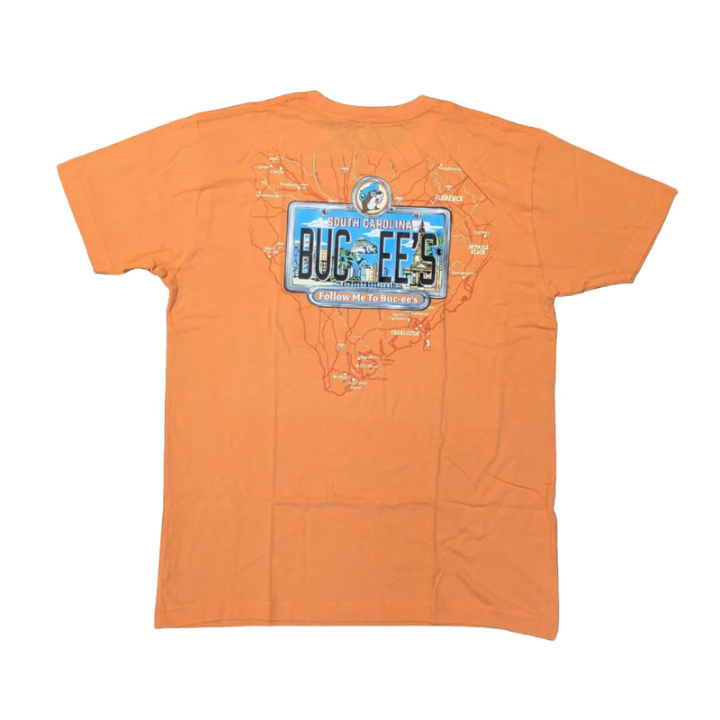 The back of an orange short-sleeve crew neck t-shirt laid flat on a white surface. The back features a large graphic of a red line-art map of South Carolina marking cities like Florence and Charleston. Overlaid on the center of the map is a blue and white custom license plate that reads "BUC-EE'S" filled with a beach cityscape illustration and the slogan "Follow Me To Buc-ee's."