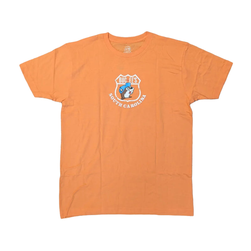 An orange short-sleeve crew neck t-shirt laid flat on a white surface. The front of the shirt features a small, centered chest graphic showing a white highway shield outline containing the Buc-ee's beaver mascot in a blue baseball cap, with the text "Buc-ee's South Carolina" curved beneath it.