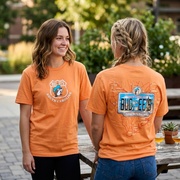 A lifestyle photograph of two women outdoors, both wearing matching orange Buc-ee's South Carolina t-shirts. One woman stands facing the camera, smiling, clearly displaying the small shield logo with the teal-capped beaver on the chest. The other woman has her back to the camera, standing next to a weathered picnic table with beer glasses, clearly showing the large detailed back graphic: a map of South Carolina overlaid with a custom license plate reading "BUC-EE'S" and the beach skyline.