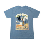 A blue slate short-sleeve crew neck t-shirt laid flat on a white background. The shirt features a large, rectangular vintage-style graphic on the chest. The design shows a beach scene with a prominent palm tree on the left, ocean waves, and a wooden signpost pointing to "Beaver Nuggets" and "Clean Restrooms." A circular logo of the Buc-ee's beaver mascot wearing a green cap is positioned near the top, bordered by the text "Buc-ee's" above and "South Carolina" along the bottom in a stylized font.