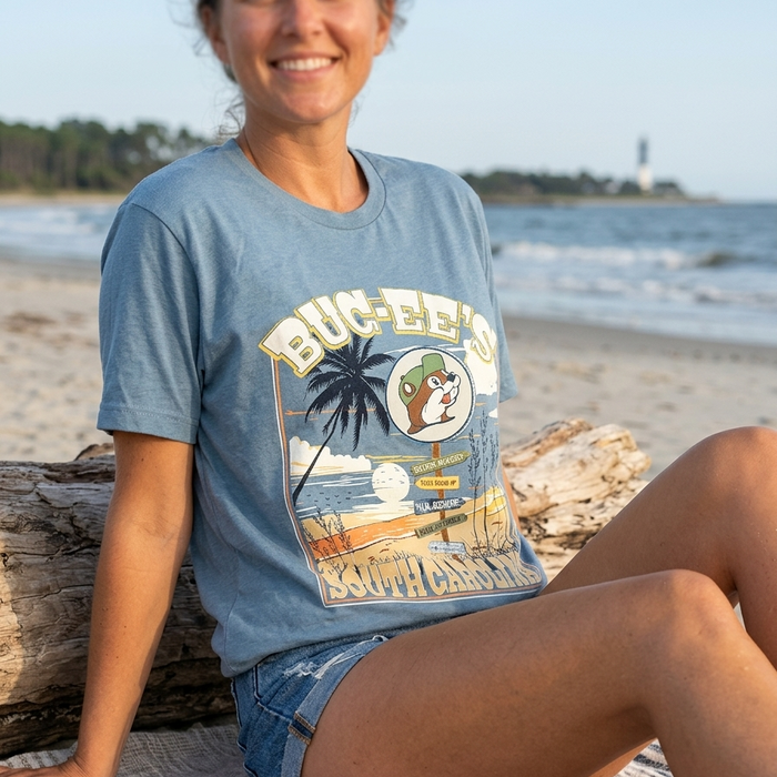 A lifestyle photograph of a smiling woman sitting on a beach blanket next to a piece of driftwood on the shore. She is wearing the blue slate Buc-ee's South Carolina t-shirt and denim shorts. The large graphic on the front of her shirt—featuring the beach signpost, palm tree, and Buc-ee's beaver mascot—is clearly visible against the ocean and distant lighthouse background.