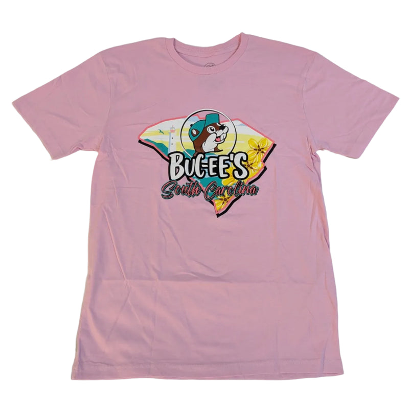 A pastel pink short-sleeve crew neck t-shirt laid flat against a white background. The shirt features a large graphic on the front showing the outline of the state of South Carolina. Inside the state shape is a retro beach sunset with a lighthouse, palm trees, and yellow flowers. Superimposed over the map is the text "Buc-ee's South Carolina" beneath a circular logo of the Buc-ee's beaver mascot wearing a teal baseball cap.