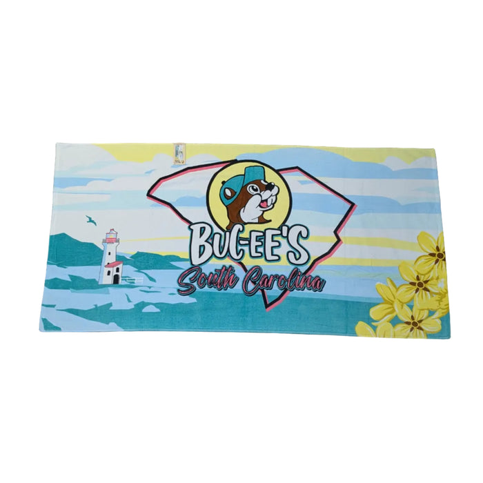 A large rectangular beach towel laid flat on a white background. The towel features a coastal design with horizontal background bands of light blue, white, and yellow. On the left side, a white lighthouse stands near a blue ocean scene. In the center, a large silhouette outline of the state of South Carolina houses a circular logo of the Buc-ee's beaver mascot wearing a blue cap, with the stylized text "Buc-ee's South Carolina" written below it. Bright yellow flowers are clustered in the lower right corner.