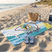 A lifestyle photograph features a Buc-ee's South Carolina beach towel spread out on the sand of a sunny beach. The towel is accented with a pair of striped flip-flops, an open book, sunglasses, a water bottle, a woven tote bag, and a cooler nearby, with the ocean waves rolling in the background.
