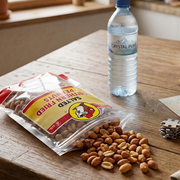 An open bag of Buc-ee's Salted Southern Fried Peanuts lies flat on a rustic wooden table, with peanuts spilling out onto the surface. Next to the bag sits a clear plastic bottle of water, a partially completed jigsaw puzzle, and a board game box.