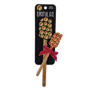 A retail package containing two Buc-ee's baking spatulas with natural wood handles, crossed over each other. A decorative red fabric ribbon with white polka dots is tied in a bow around the handle of the smaller spatula. The large spatula has a black silicone head and the smaller one has a red head, both covered in a repeating yellow Buc-ee's beaver logo print.