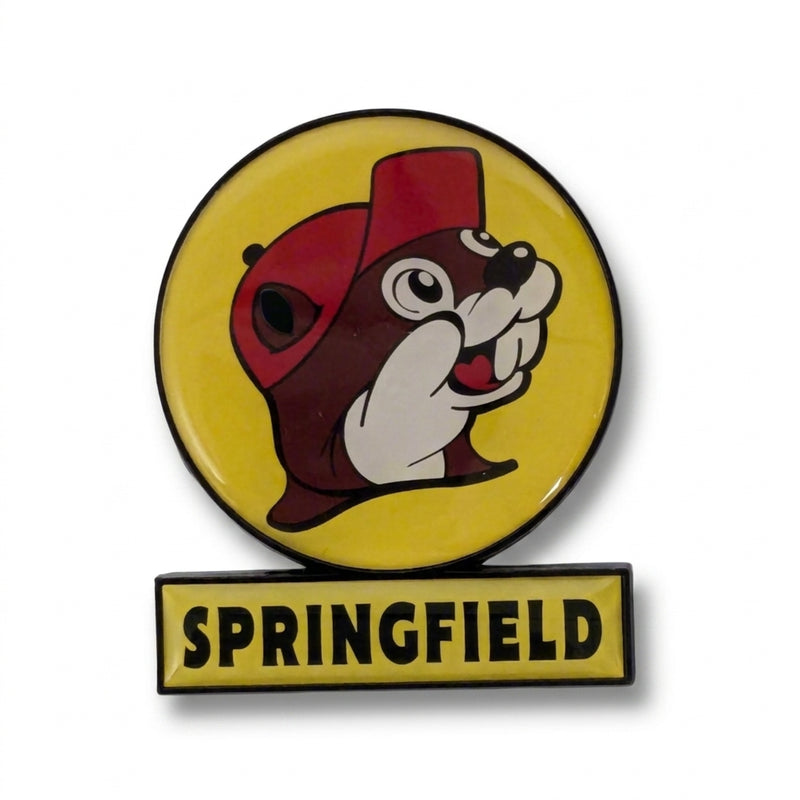 A circular yellow souvenir fridge magnet shaped like a Buc-ee's highway billboard sign on a solid white background. It features the smiling beaver mascot logo in a red cap inside a yellow circle, mounted over a rectangular yellow bar that reads "SPRINGFIELD" in bold black lettering.
