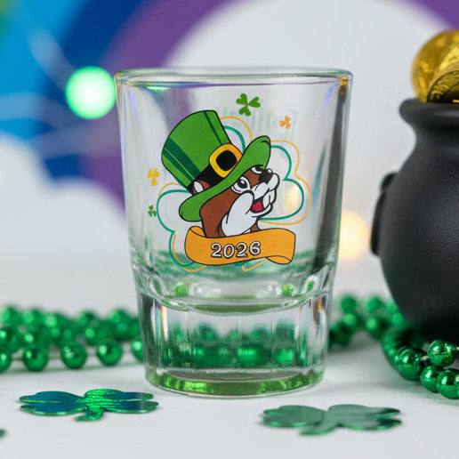 A clear glass shot glass with a green base. The front features the Buc-ee's beaver mascot wearing a green leprechaun hat and a yellow bow tie, with the year "2026" printed on a golden banner below him. The shot glass is placed beside a small black pot filled with gold chocolate coins with a rainbow backdrop effect, green beads, and clover decorations.