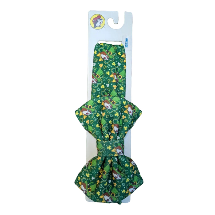 A Buc-ee's green headband with a green bow with a dense pattern of festive beavers and colorful clovers, mounted on a white retail display card with the Buc-ee's logo, all displayed on a clean white background.