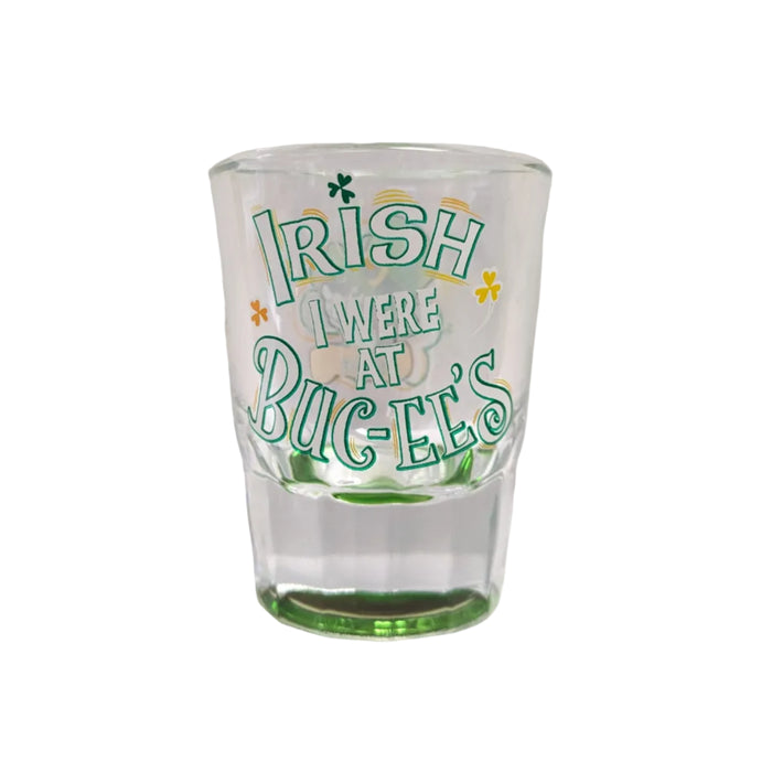 The back of a clear shot glass featuring the text "IRISH I WERE AT BUC-EE'S" in stylized green and white letters, decorated with small green and orange shamrocks, displayed on a clean white background.
