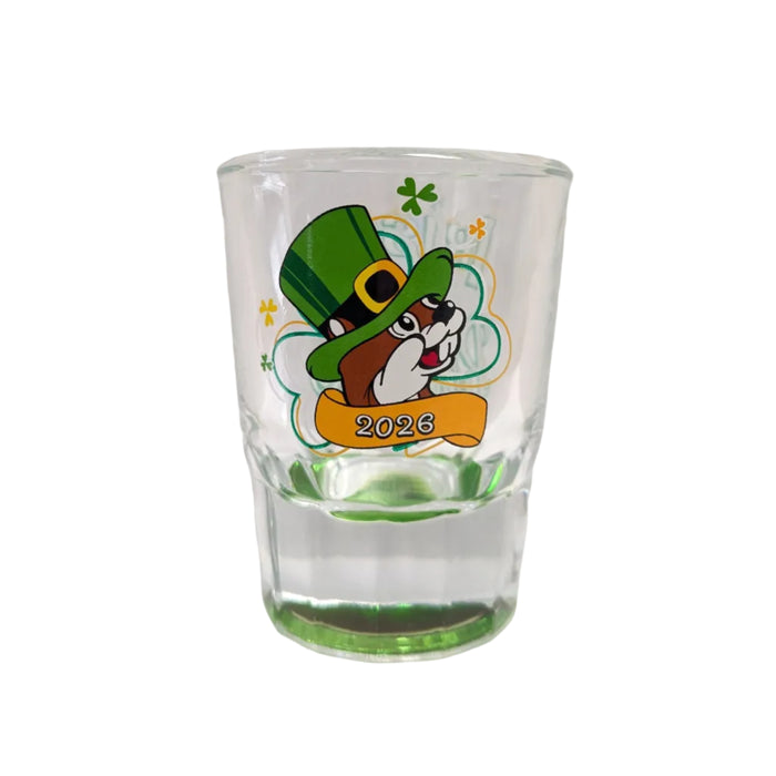 A clear glass shot glass with a green base. The front features the Buc-ee's beaver mascot wearing a green leprechaun hat and a yellow bow tie, with the year "2026" printed on a golden banner below him, displayed on a clean white background.