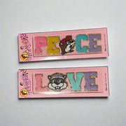 Two Buc-ee’s sticker patches shown on pink packaging: one spells “LOVE” with Buc-ee’s face as the “O,” and the other spells “PEACE” with Buc-ee in the center, both in colorful glitter letters.

