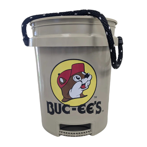 A front-facing view of a tan 5-gallon plastic bucket featuring the circular yellow and red Buc-ee’s beaver logo. The bucket includes a thick black-and-white braided rope handle and a specialized molded handle at the bottom base designed to assist with controlled pouring. Displayed on a clean white background.
