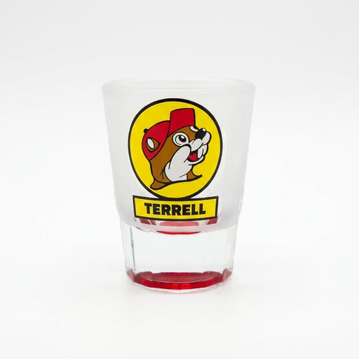 A clear frosted shot glass featuring the yellow circular Buc-ee's beaver logo with a yellow banner underneath reading "TERRELL" in black text, set against a solid white background. The glass features a thick clear base tinted red at the very bottom.