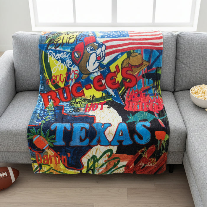 Buc-ee’s Texas plush blanket with colorful collage design, American flag, Buc-ee the Beaver graphic, and large “TEXAS” lettering laid on the front of a couch with a football on the floor and a bowl of popcorn next to it.