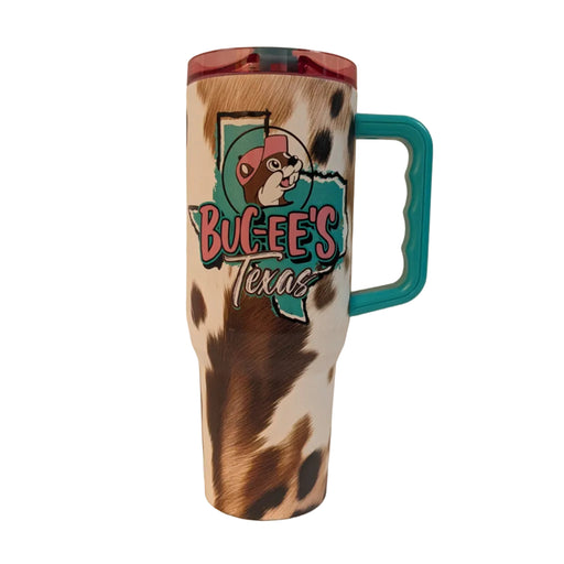 A 40 oz insulated travel tumbler with a brown and white cowhide pattern print. The front features a teal silhouette of the state of Texas with a black outline, layered with the words "Buc-ee's Texas" and the Buc-ee's beaver mascot wearing a pink baseball cap. The tumbler includes a bright teal plastic handle on the side and a translucent pink lid.