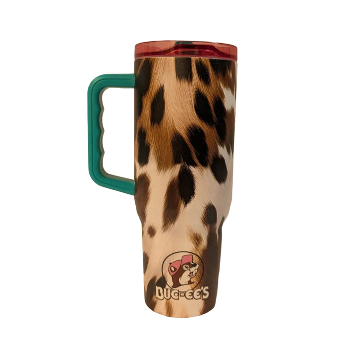 A rear view of the 40 oz insulated travel tumbler, showcasing the continuation of the brown, black, and white cowhide pattern print. A smaller, circular Buc-ee's logo featuring the beaver mascot wearing a pink baseball cap is centered near the tapered base with "BUC-EE'S" written in white block letters below it. The bright teal plastic handle extends from the left side, and the translucent pink lid is visible at the top.