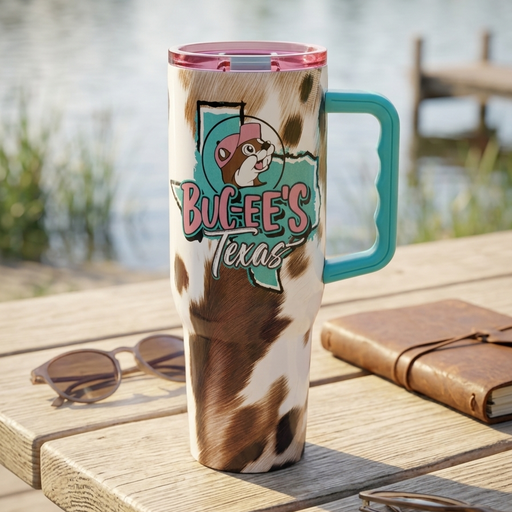 A lifestyle photograph of a Buc-ee's 40 oz insulated travel tumbler standing upright on a weathered wooden picnic table. The tumbler features a bold brown, black, and white cowhide pattern and a solid teal graphic of the state of Texas with a black outline. Layered over the state graphic is the Buc-ee's beaver mascot wearing a pink baseball cap and the text "Buc-ee's Texas." The tumbler has a prominent teal side handle and a translucent pink lid.