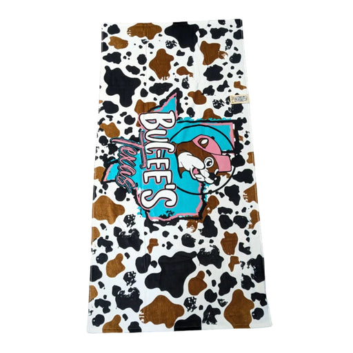 A playful beach towel with a brown and white cow-hide print background. In the center, a turquoise silhouette of the state of Texas is overlaid with the "Buc-ee's" name in white and "Texas" in a pink western font. Displayed on a clean white background.