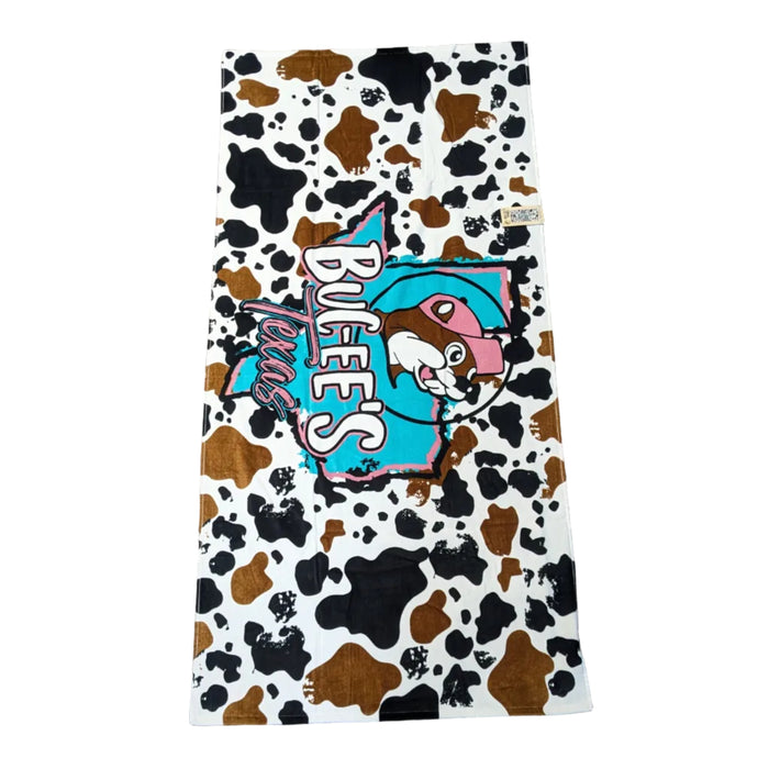 A playful beach towel with a brown and white cow-hide print background. In the center, a turquoise silhouette of the state of Texas is overlaid with the "Buc-ee's" name in white and "Texas" in a pink western font. Displayed on a clean white background.