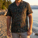 Man wearing a patterned shirt standing on a beach with water and trees in the background