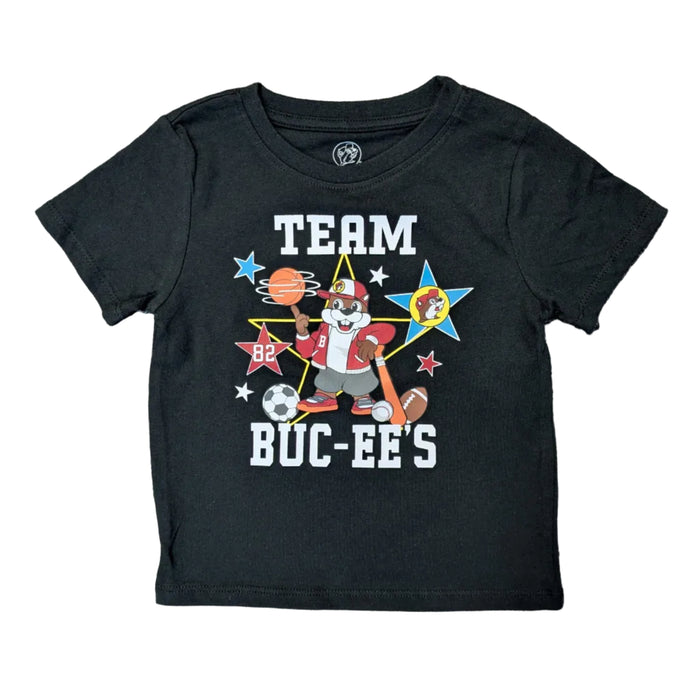 A black toddler-sized t-shirt laid flat on a white background. The shirt features a colorful graphic in the center with the words 'TEAM BUC-EE'S' in bold white letters. The graphic shows the Buc-ee's beaver mascot wearing a red varsity jacket and holding a baseball bat, surrounded by a basketball, soccer ball, football, and various blue, red, and white stars.