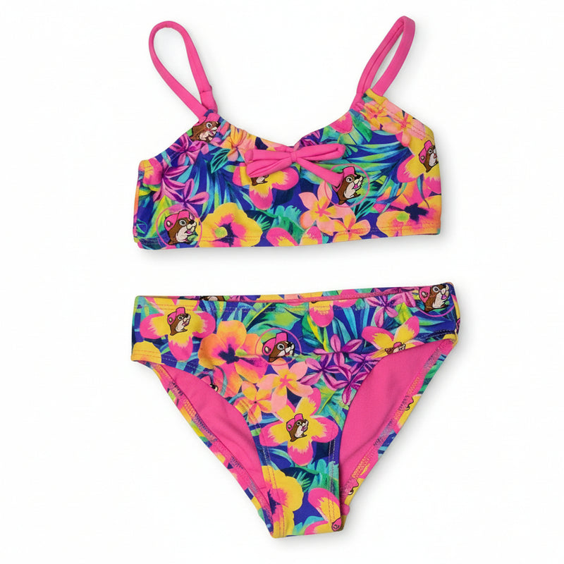 A Buc-ee's girls' two-piece bikini featuring a vibrant tropical floral print with pink and yellow hibiscus flowers and the Buc-ee's mascot, styled with thin pink straps, a decorative center bow on the top, and a solid pink interior lining, displayed on a clean white background.