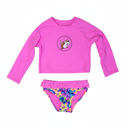 A two-piece swim set for infants and toddlers featuring a solid bright pink long-sleeve rash guard top with a circular graphic of the Buc-ee's mascot in a pink hat centered on the chest. The matching swim bottoms feature a vibrant tropical floral print with hibiscus flowers and the Buc-ee's mascot, finished with a ruffled pink waistband and a solid pink interior lining, displayed on a clean white background.