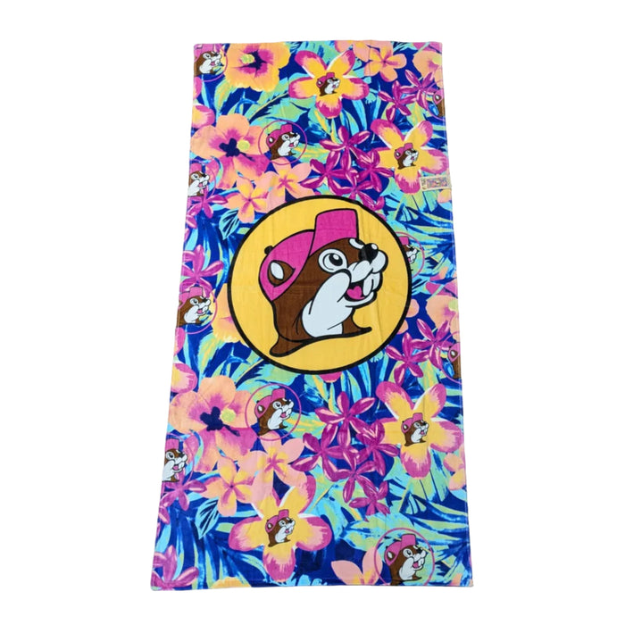A bright beach towel with a dense tropical pattern of pink hibiscus flowers and orange blossoms over a leafy dark blue and teal background. A yellow circle with a black border in the middle  with the iconic Buc-ee's beaver mascot wearing a pink hat. Displayed on a clean white background.