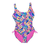 A Buc-ee's Women's one-piece tropical floral swimsuit featuring a vibrant tropical floral print with pink and yellow hibiscus flowers and the Buc-ee's mascot, styled with cinched sides and side tie detail, tied in bows above the leg holes, and a solid pink interior lining, displayed on a clean white background.