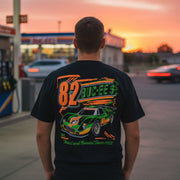 Man standing at a gas station wearing a black Buc-ee’s Turnin’ and Burnin’ t-shirt, shown from the back with a colorful racing graphic and Buc-ee’s beaver design.