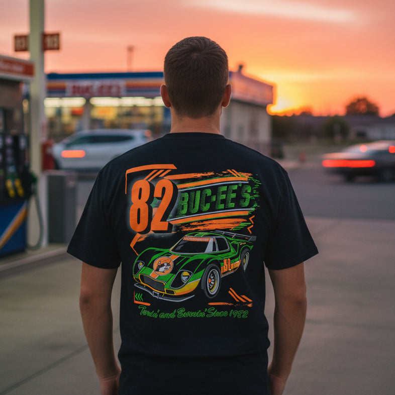 Man standing at a gas station wearing a black Buc-ee’s Turnin’ and Burnin’ t-shirt, shown from the back with a colorful racing graphic and Buc-ee’s beaver design.
