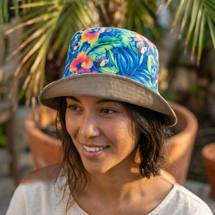 An AI-generated lifestyle photograph of a woman wearing the reversible Buc-ee’s bucket hat in a sunny outdoor setting. The hat is worn with the tropical side facing out, featuring a vibrant blue, teal, and orange hibiscus floral pattern with the Buc-ee's beaver mascot integrated into the design. The brim is slightly upturned, revealing the solid khaki-colored underside. The woman is smiling and looking off-camera, with a blurred background of a palm tree and terracotta garden pots.