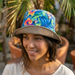 An AI-generated lifestyle photograph of a woman wearing the reversible Buc-ee’s bucket hat in a sunny outdoor setting. The hat is worn with the tropical side facing out, featuring a vibrant blue, teal, and orange hibiscus floral pattern with the Buc-ee's beaver mascot integrated into the design. The brim is slightly upturned, revealing the solid khaki-colored underside. The woman is smiling and looking off-camera, with a blurred background of a palm tree and terracotta garden pots.