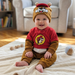 A happy, smiling baby sitting on a soft cream-colored blanket on a wooden floor in a cozy nursery. The baby is wearing a Buc-ee's themed outfit, including a red and brown fleece long-sleeved bodysuit with the iconic beaver logo on the chest and a matching brown fleece beanie that features the beaver's face and soft ears. In the background, a comfortable armchair and a bookshelf filled with colorful children's books are softly blurred.