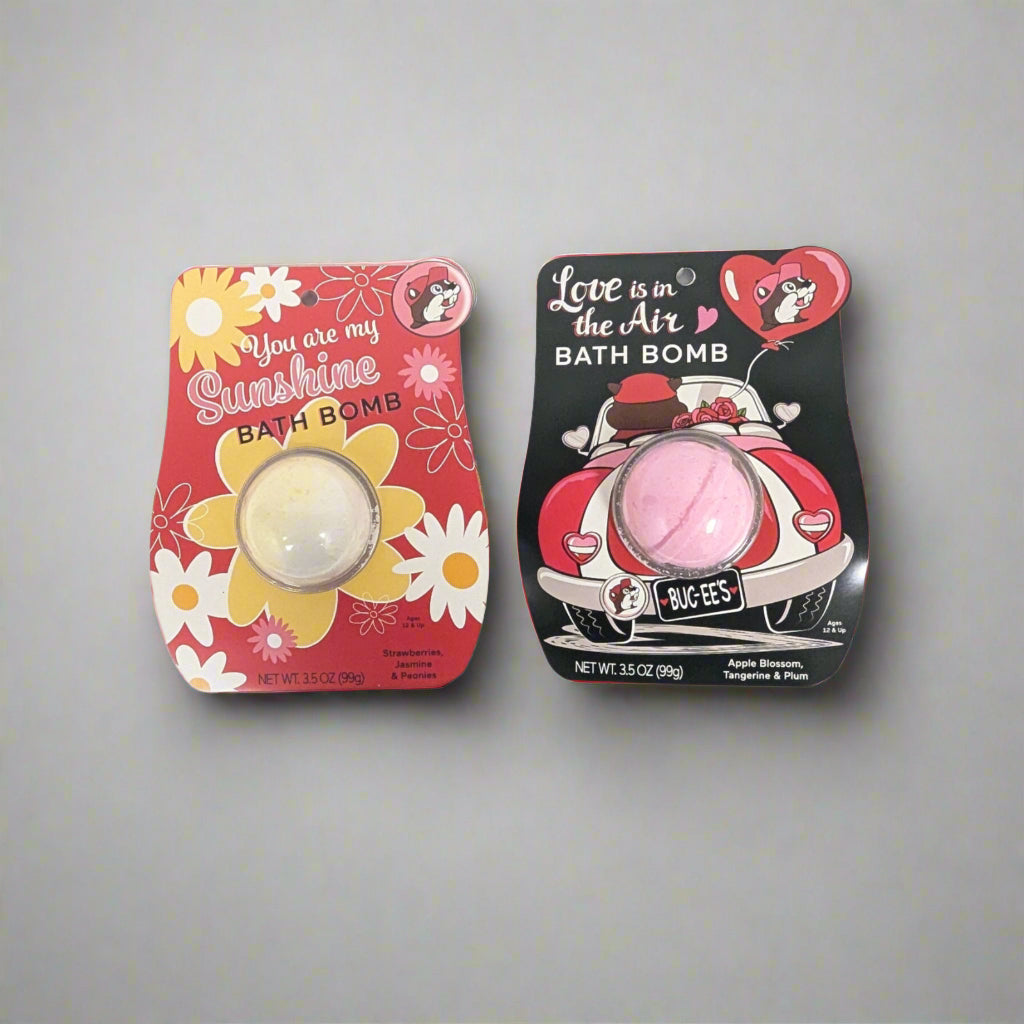 Top-down view of two Buc-ee's bath bombs. One is labeled "You Are My Sunshine" with a red floral design, and the other says "Love Is in the Air" with a pink retro car illustration.