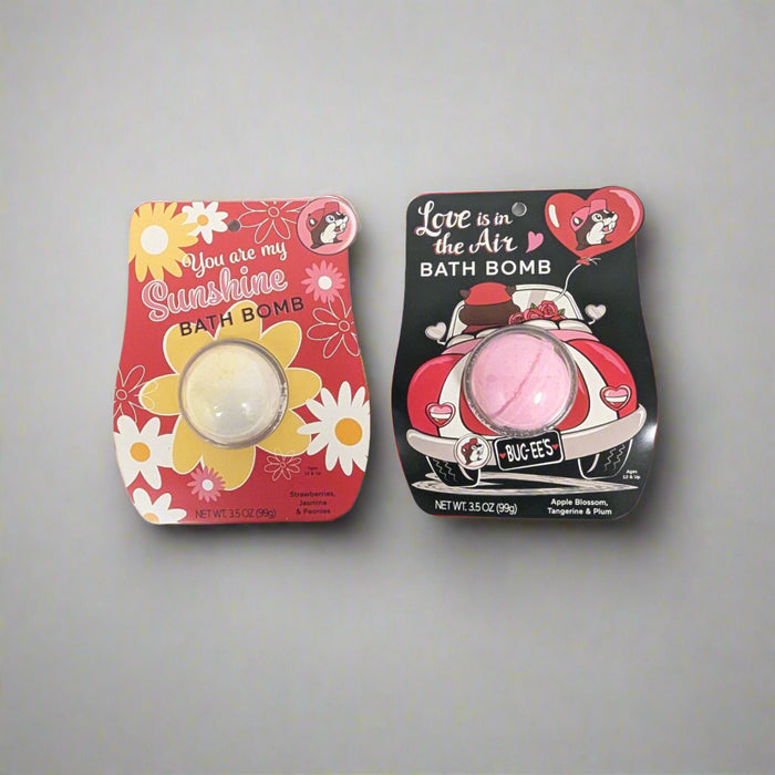 Top-down view of two Buc-ee's bath bombs. One is labeled "You Are My Sunshine" with a red floral design, and the other says "Love Is in the Air" with a pink retro car illustration.