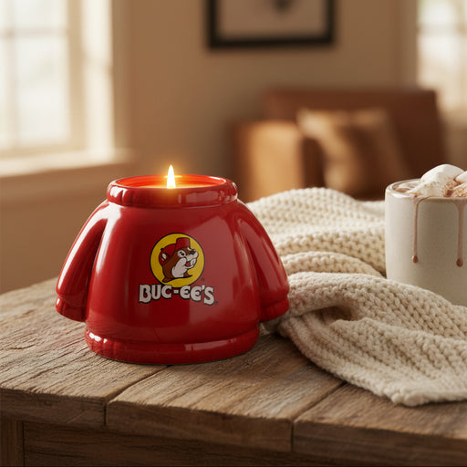 Red ceramic Buc-ee’s hoodie-shaped candle jar featuring the Buc-ee’s logo, part of the Warm Cashmere scented candle, displayed on a table with a blanket and cup of hot cocoa.